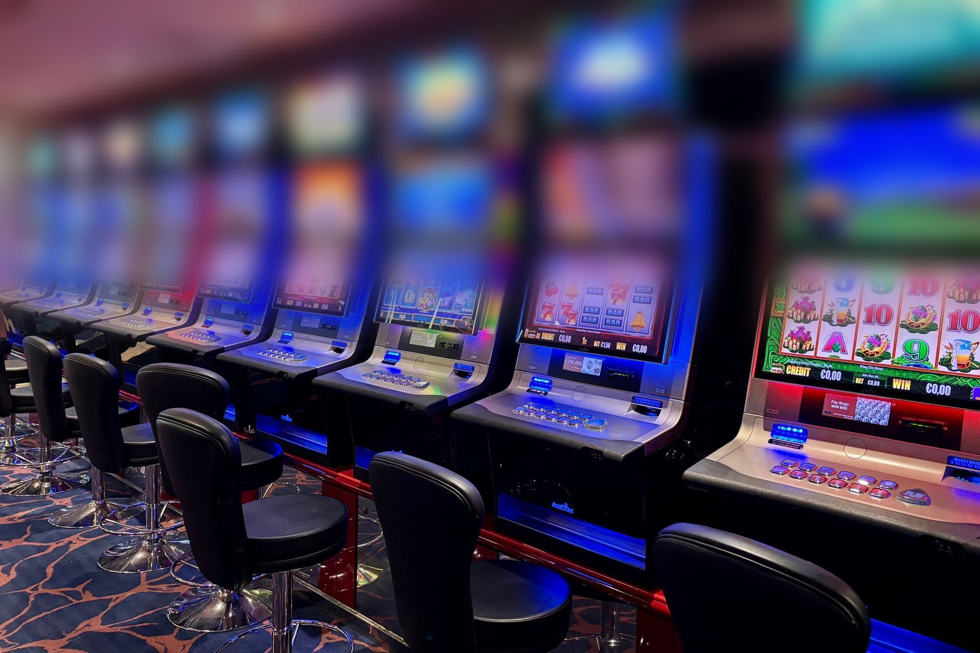 A row of empty slot machines with glowing reel screens and black stools on a casino floor