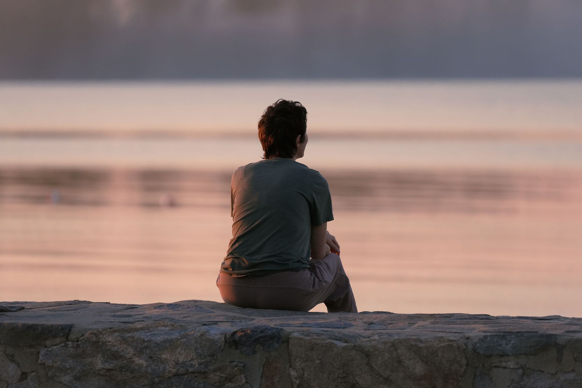 A person seen from behind sitting on a breakwall, watching a calm sunrise over the sea