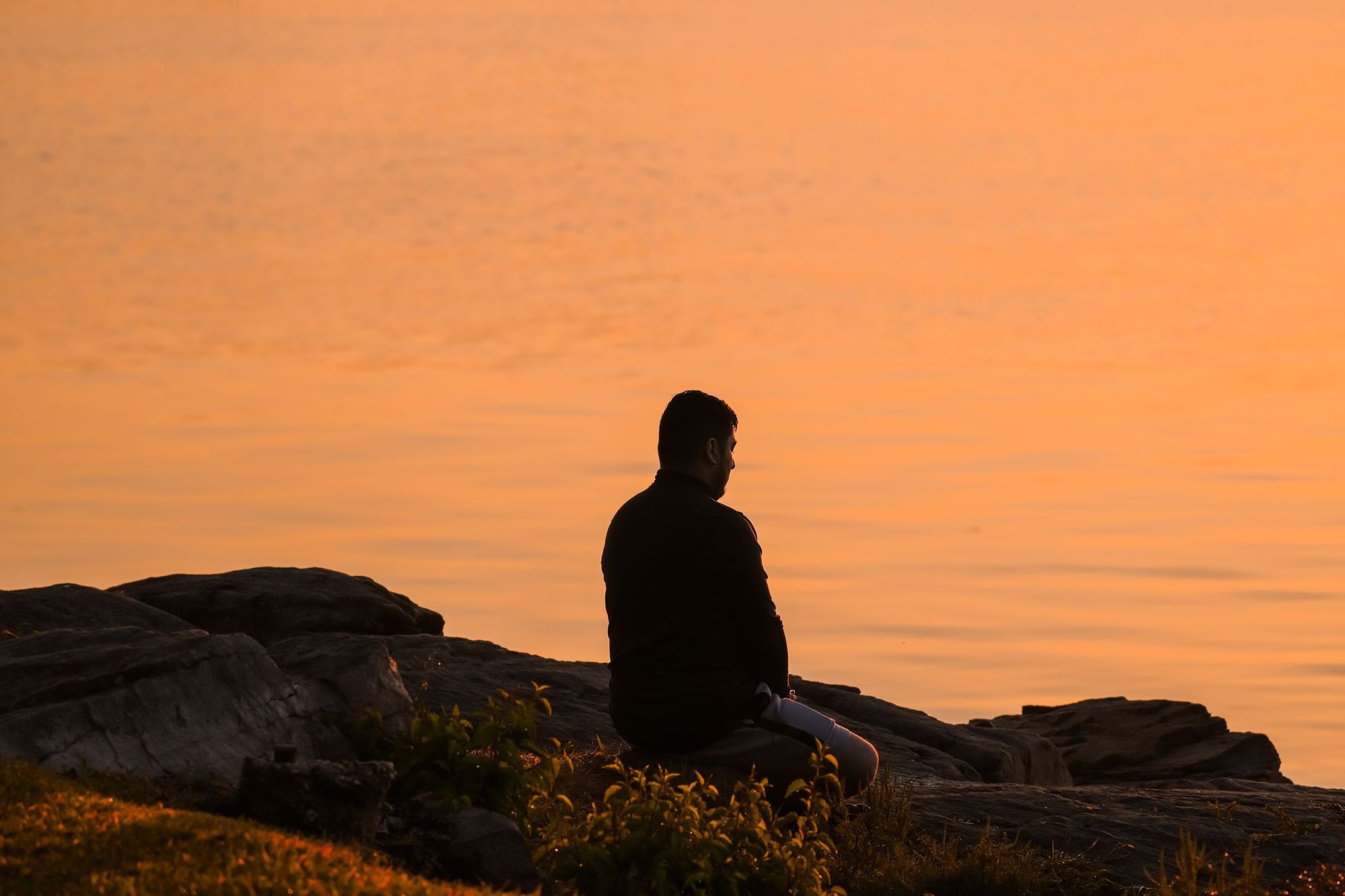 The silhouette of a person sitting quietly on rocks by the water against an orange sunrise